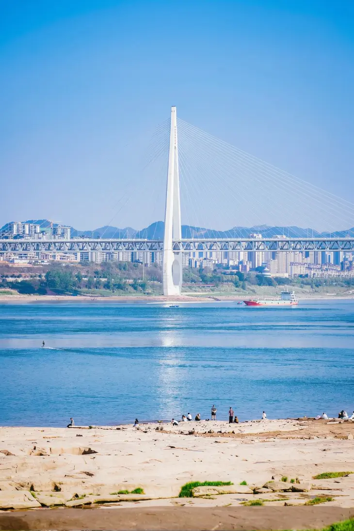 Wide view of Chongqing beach riverbank with blue water and Chaotianmen Yangtze River Bridge under a sunny sky